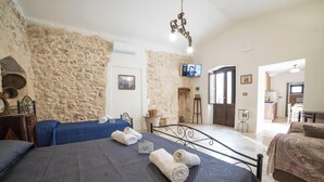 Habitación de hotel con pared de piedra, una cama con colcha azul oscuro, una segunda cama con colcha azul, un candelabro, un televisor y vista de la cocina a través de un vano abierto.