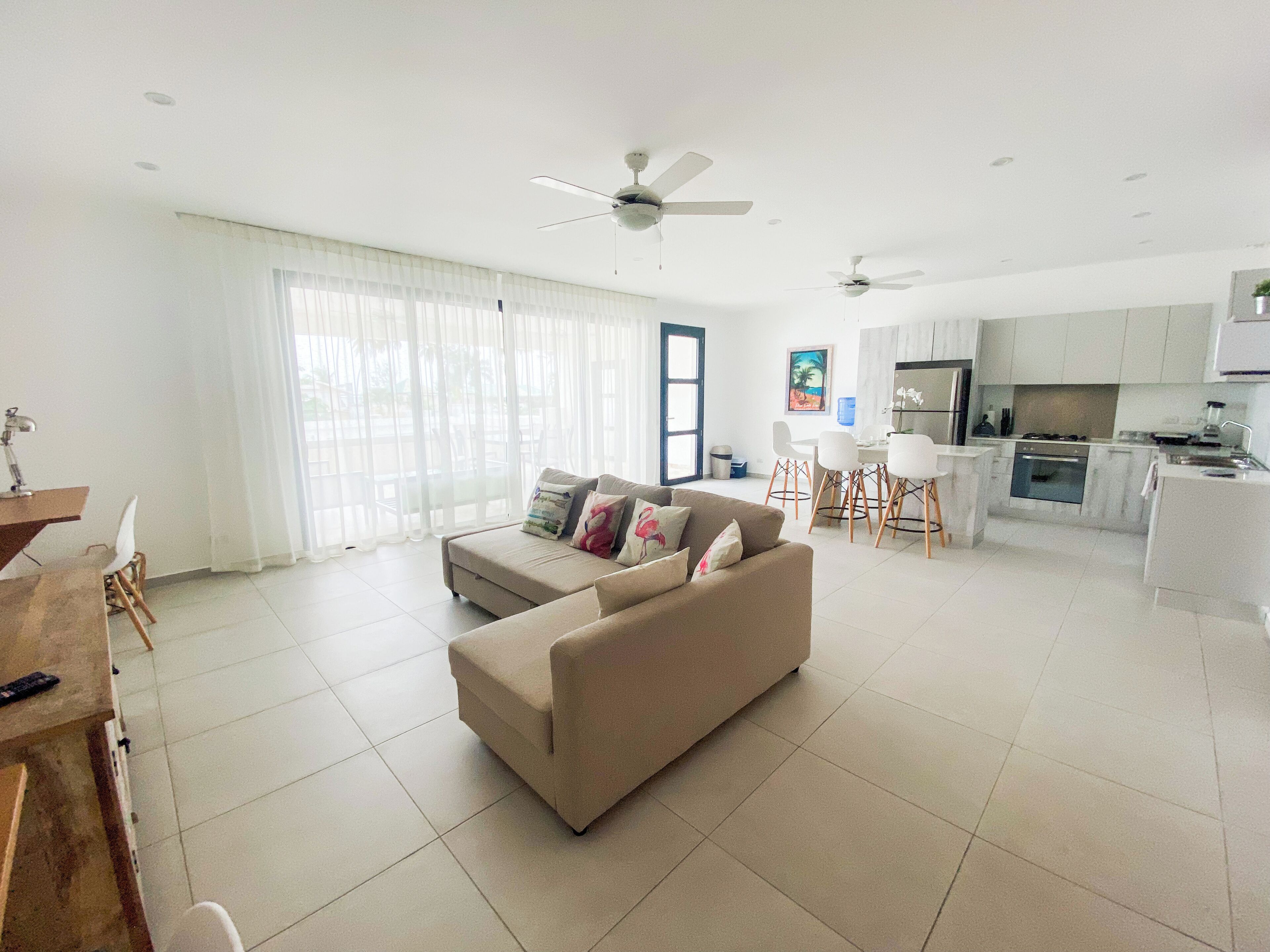 A modern living room with a beige sofa, a dining area with white chairs, and a kitchen with white cabinets.
