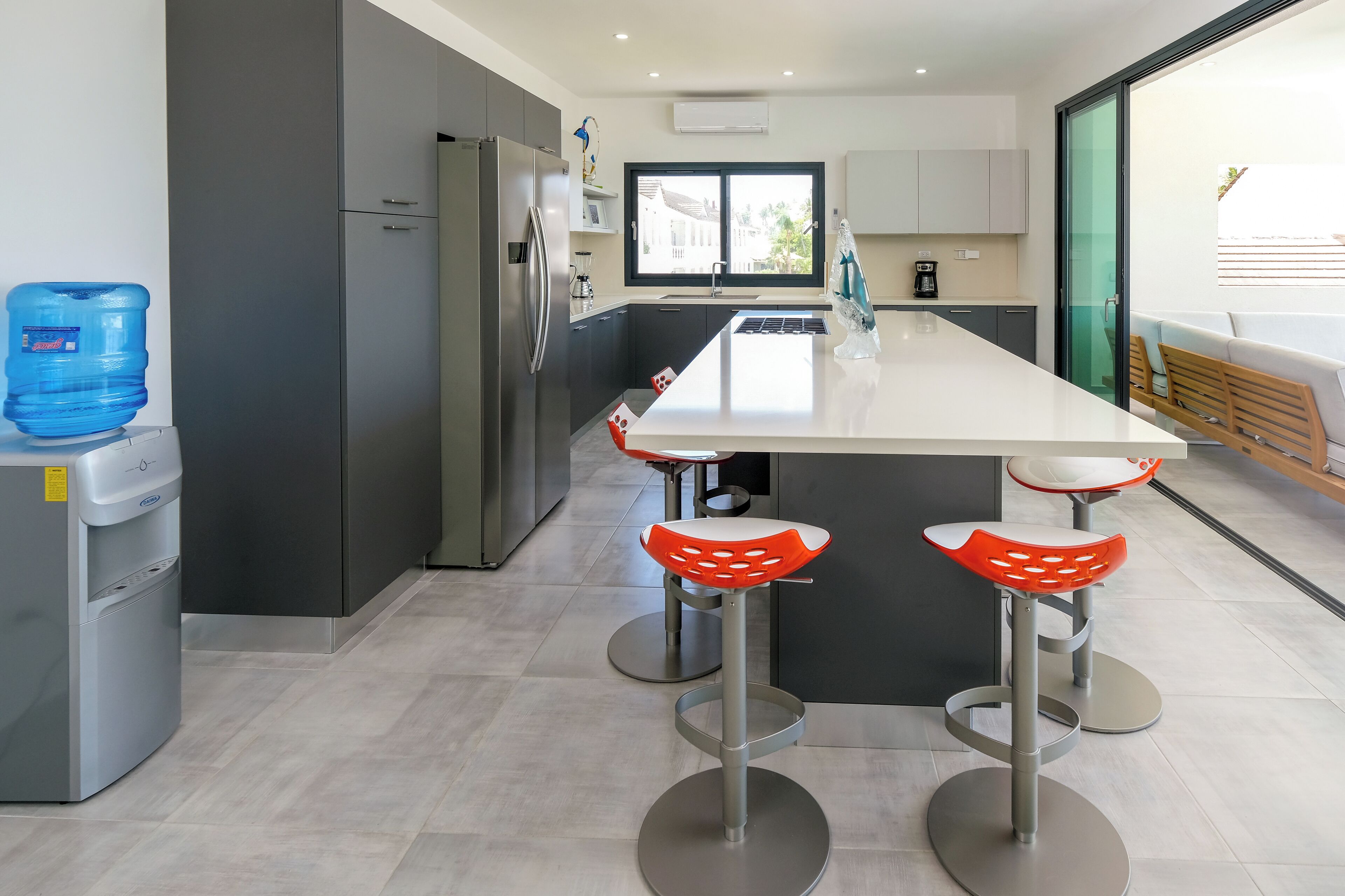 Una cocina moderna con una isla central, dos taburetes de bar, un dispensador de agua y un refrigerador.