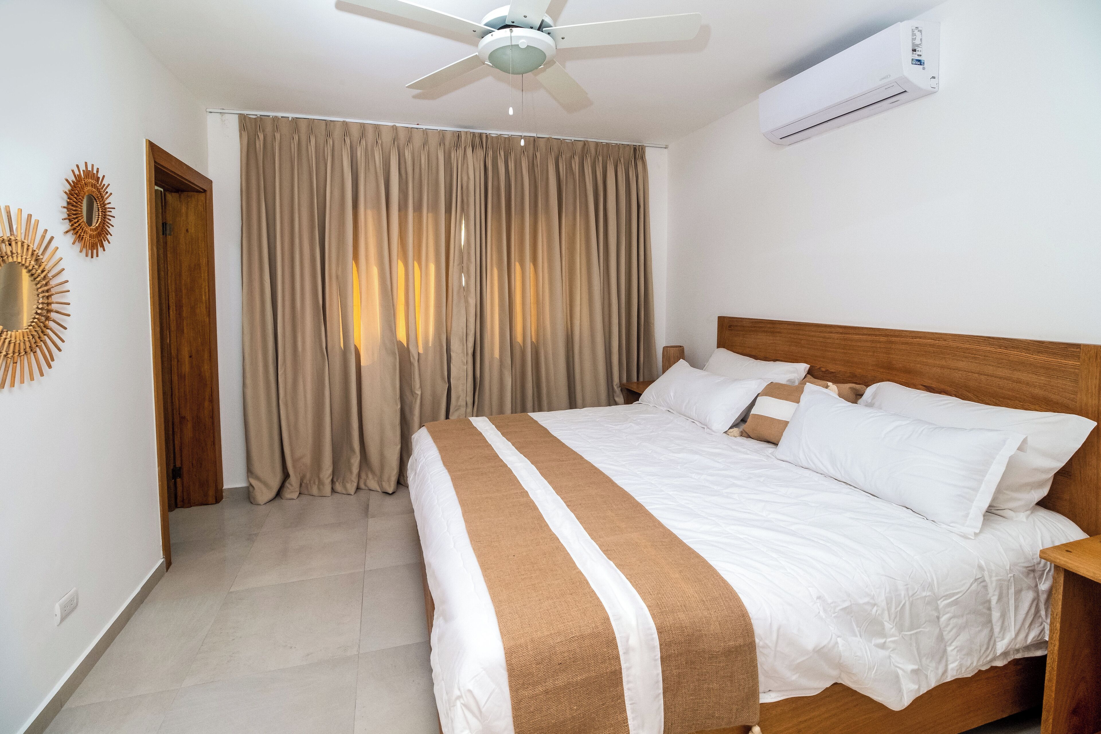 A neatly made bed with white and brown bedding, a ceiling fan, and an air conditioning unit on the wall.