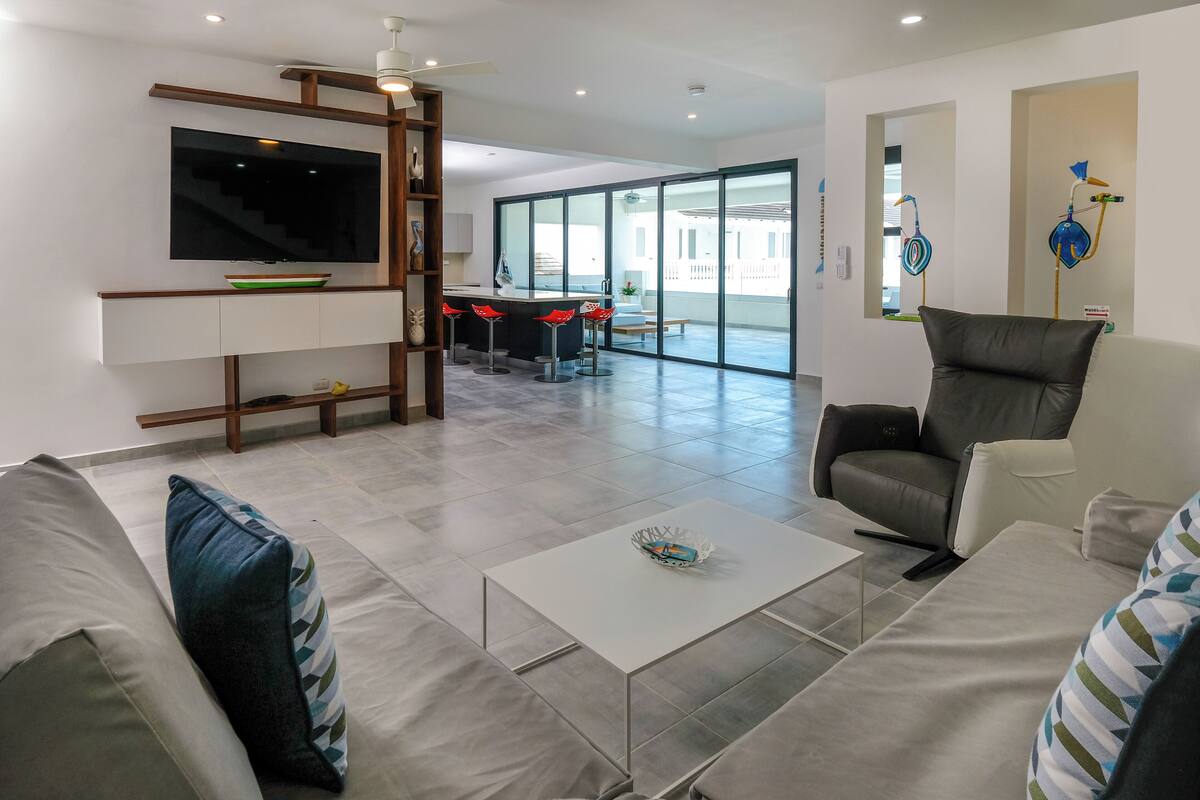 A modern living room with a flat-screen TV, a sofa, a coffee table, and a dining area with red chairs.