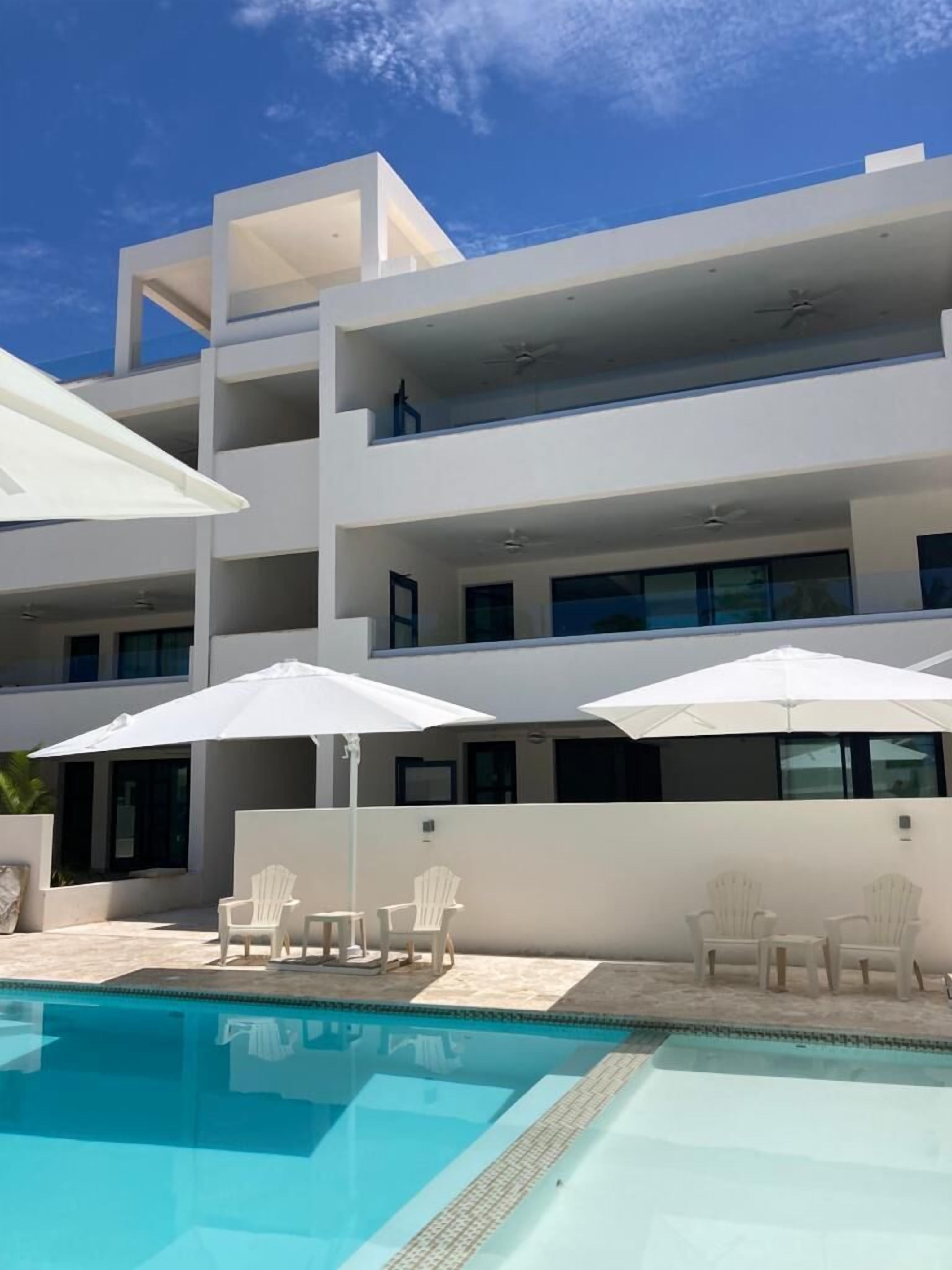 A modern multi-story building with a swimming pool, outdoor seating, and a clear blue sky.