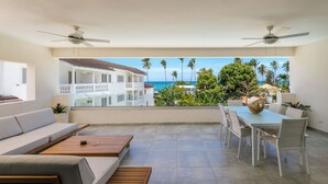 A modern living room with a balcony, white furniture, and a view of a multi-story building and palm trees. - Stella Apart-hotel Las Terrenas (Las Terrenas)