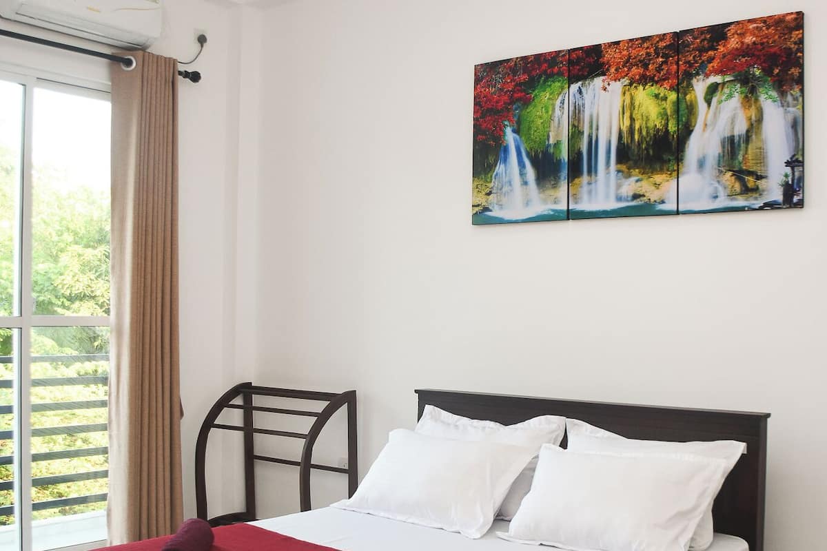 Quarto de hotel com cama, ventoinha no teto, janela com cortinas e quadro de uma cascata.