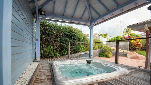 A hot tub under a covered area with a wooden deck, surrounded by greenery and a view of a residential area.
