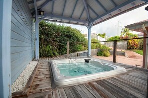 Un jacuzzi bajo un techo, con una terraza de madera, rodeado de follaje y vistas a una zona residencial.