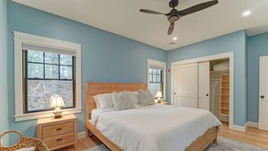 Un dormitorio con una cama de madera, una mesita de noche con lámpara, un ventilador de techo y una ventana con vista a árboles.