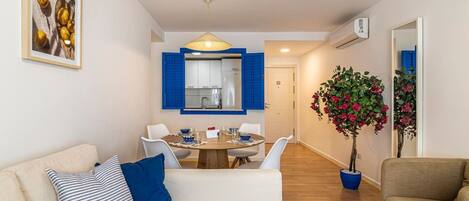 A modern living room with a sofa, a dining table with chairs, a kitchen area with blue shutters, and a potted plant.