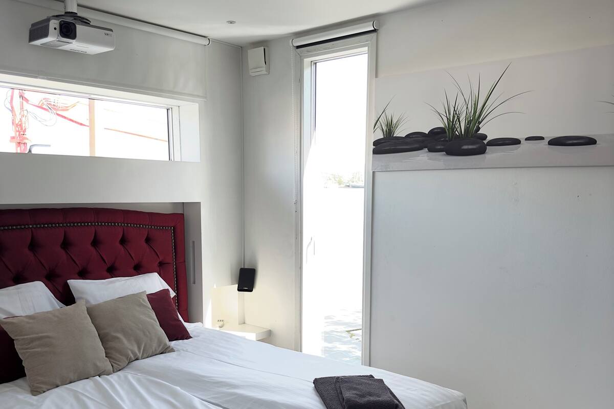 Ein Hotelzimmer mit einem großen Bett, einem roten Kopfteil, einem Deckenprojektor, einem Fenster mit Jalousien, einem Nachttisch, einer Wand-Klimaanlage und einem kleinen Topfpflanzen auf der Fensterbank.