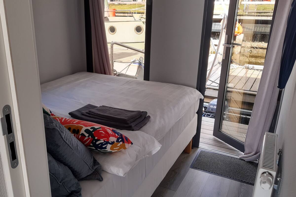 Ein kleines, ordentlich bezogenes Bett mit farbigem Kissen und gefaltetem Bettbezug, ein Fenster mit Ausblick und eine Tür zu einem Außenbereich.