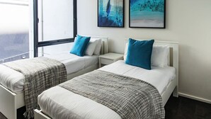 Ein Hotelzimmer mit zwei Betten, jeweils mit weißer Matratze und blauen Kissen, einer grauen Decke und einem kleinen Nachttisch. Darüber an der Wand befinden sich zwei gerahmte Kunstwerke.