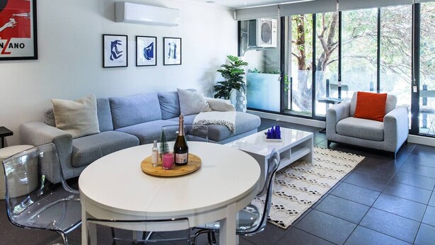 Ein modernes Wohnzimmer mit einem grauen Sofa, einem runden Couchtisch mit einer Flasche und einem großen Fenster mit Blick auf Bäume.