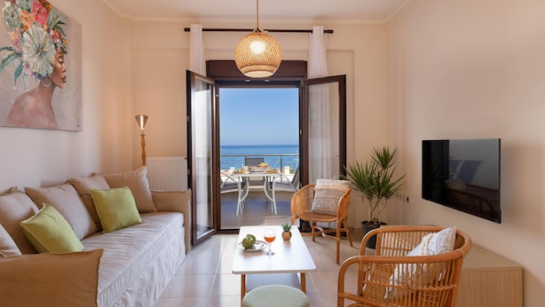 A modern living room with a sofa, chairs, a table with fruit and drinks, a TV, and a view of the sea through an open door. - Green A Seaside Escape With Mesmerizing Views (Rethymno)