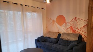 Un salon avec un canapé bleu foncé, deux tables rondes et une fresque murale représentant des motifs abstraits de montagnes et de soleils.