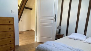 Een slaapkamer met een houten dressoir, een bed met wit beddengoed, een nachtkastje met een lamp en een deur naar een andere kamer.