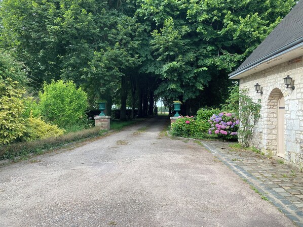 En grusvej fører hen til en bygning i sten med sokkel af mursten og rundbueindgang.
