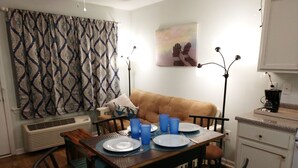 A dining area with a table set for four, a brown sofa, patterned curtains, and a painting of chairs on the wall.