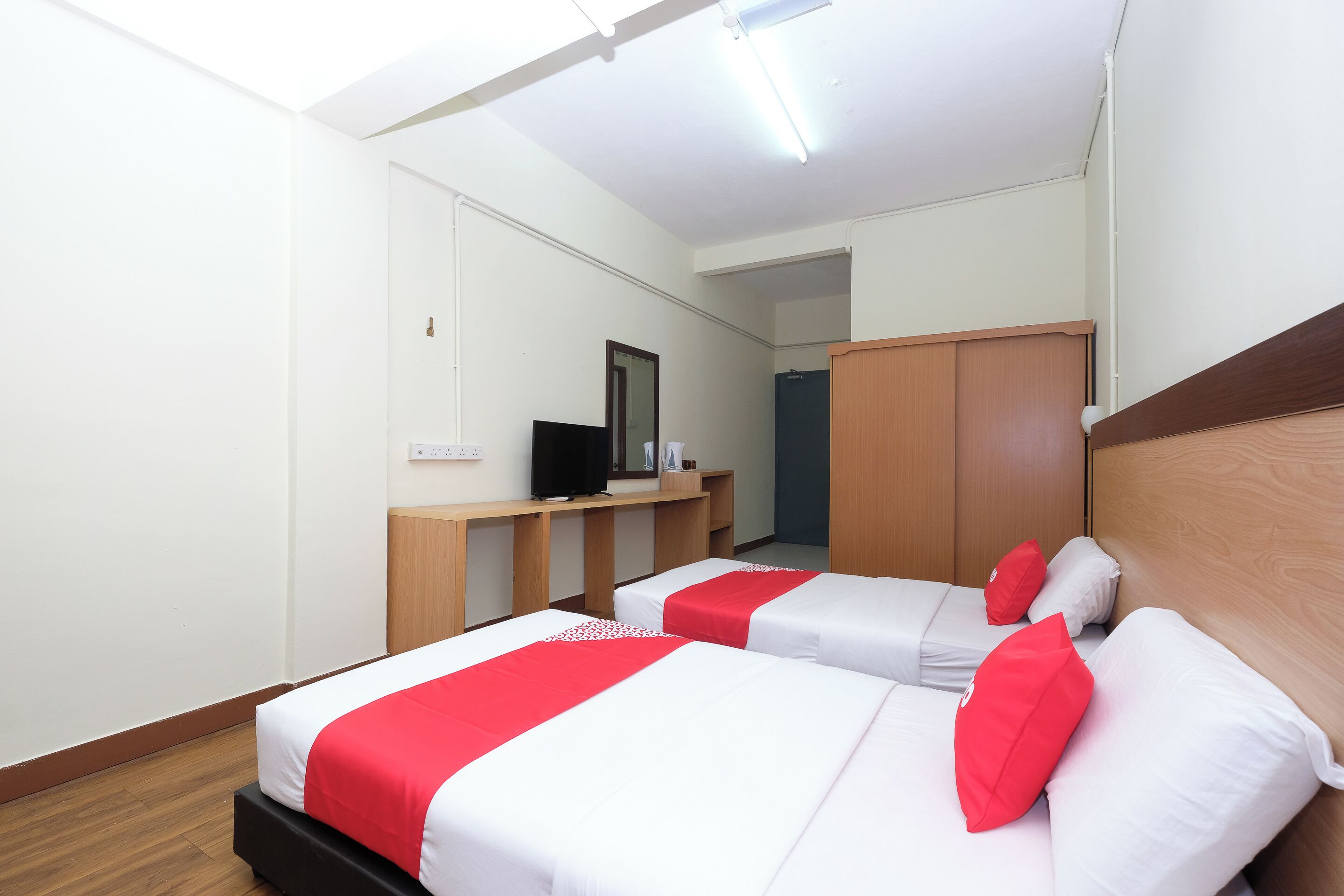 A hotel room with two beds, each with red and white bedding, a wooden headboard, a desk with a computer, and a mirror on the wall.