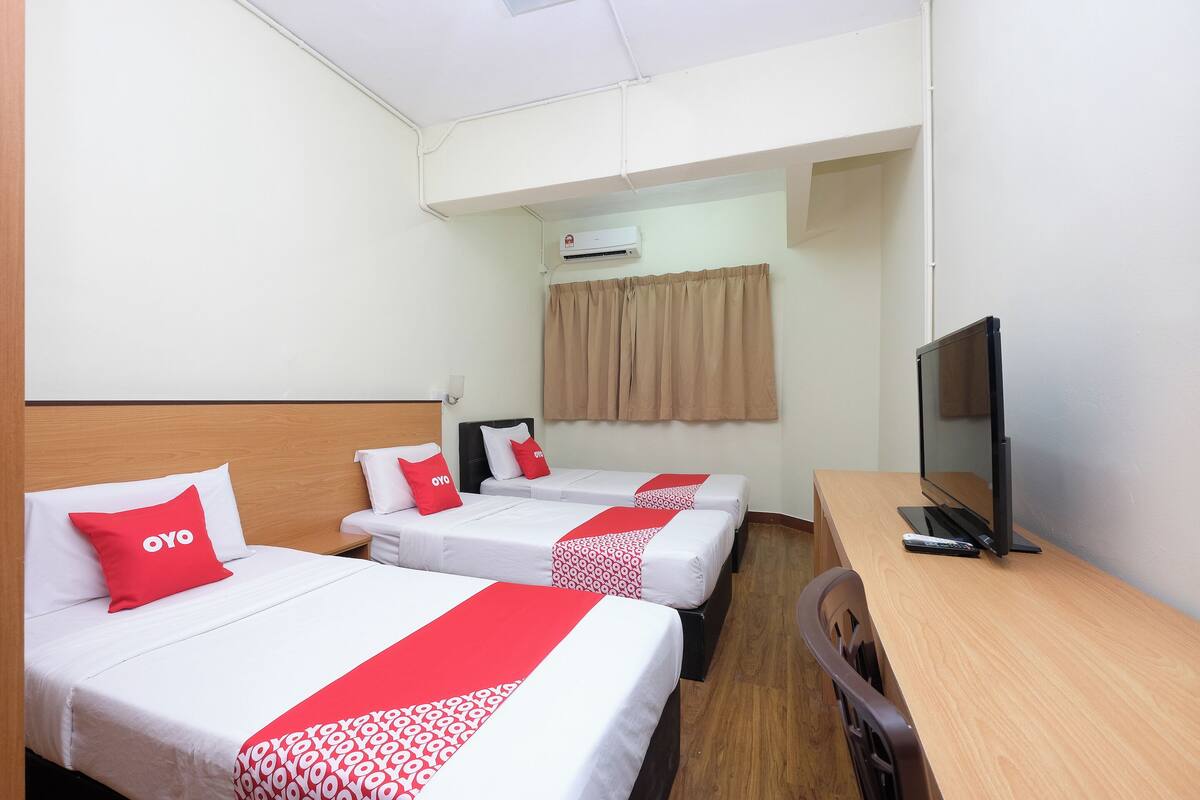A hotel room with three beds, a desk, a television, and an air conditioning unit.