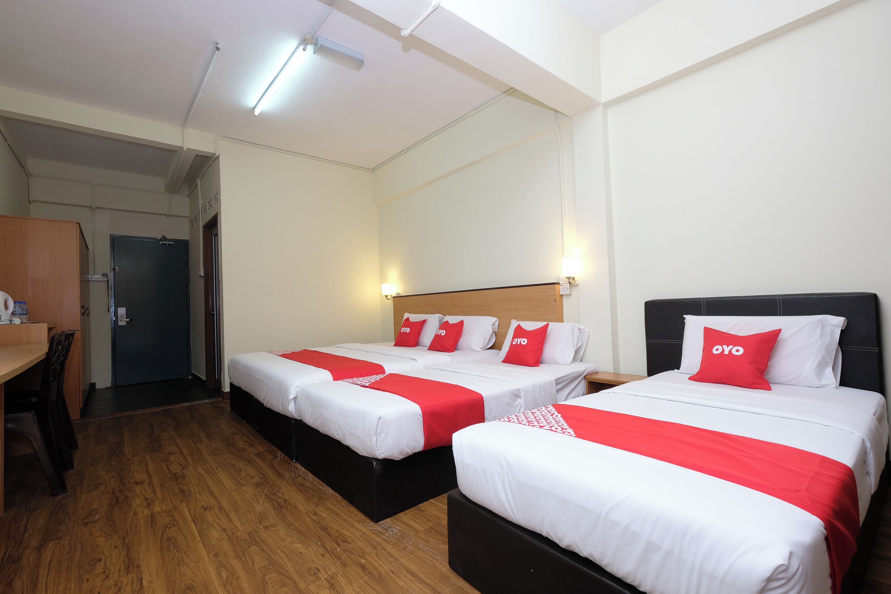 Quarto de hotel com duas camas, cada uma com roupa de cama vermelha e branca, uma escrivaninha de madeira e uma porta ao fundo.