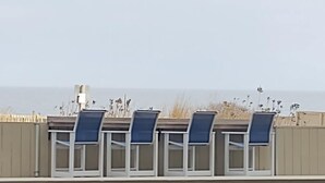 A row of blue chairs with white frames, aligned against a beige wall, overlooking a beach with tall grasses and the sea in the background. - Cozy Studio l Oceanfront l Pool l WiFi (Ocean City)