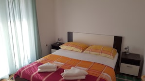 Ein ordentlich bezogenes Bett mit einer Karo-Decke und zwei Kissen, ein Nachttisch mit Lampe sowie ein Beistelltisch mit Lampe.