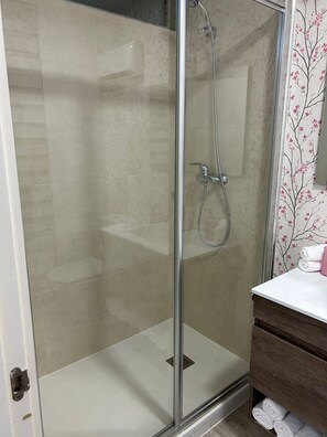 Une douche fermée par des parois en verre, dotée d’un pommeau de douche à main, un lavabo blanc avec un meuble-vasque en bois et un porte-serviettes avec des serviettes pliées.