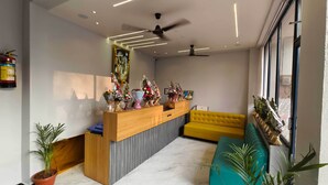 A reception area with a wooden desk, a yellow sofa, and potted plants. - Hotel Shridhar (Ujjain)
