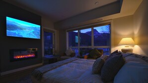 Een slaapkamer met een groot bed, een aan de muur bevestigde televisie, een open haard en uitzicht op bergen door het raam.