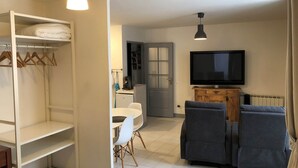 A modern living room with a flat-screen TV, two grey sofas, a dining table with chairs, a white wardrobe, and a small kitchenette.