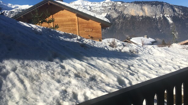 Una cabaña de madera con techo nevado, con montañas y cielo azul despejado al fondo.
