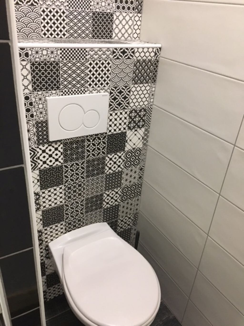 Une salle de bain équipée de toilettes, d’une chasse d’eau murale et d’un mur carrelé orné de différents motifs géométriques.