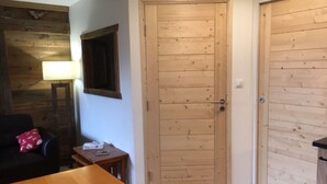 Una habitación con puerta de madera, una mesita pequeña, una lámpara y un sofá negro con un cojín rojo.