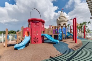 A playground with slides and climbing structures, surrounded by a fenced area and palm trees.