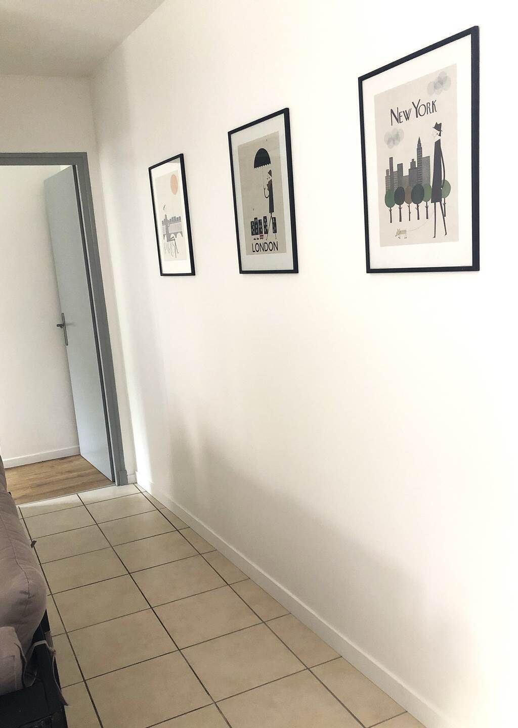 Un couloir avec trois tableaux encadrés représentant des paysages urbains accroch és au mur, une porte au fond et un sol carrelé.