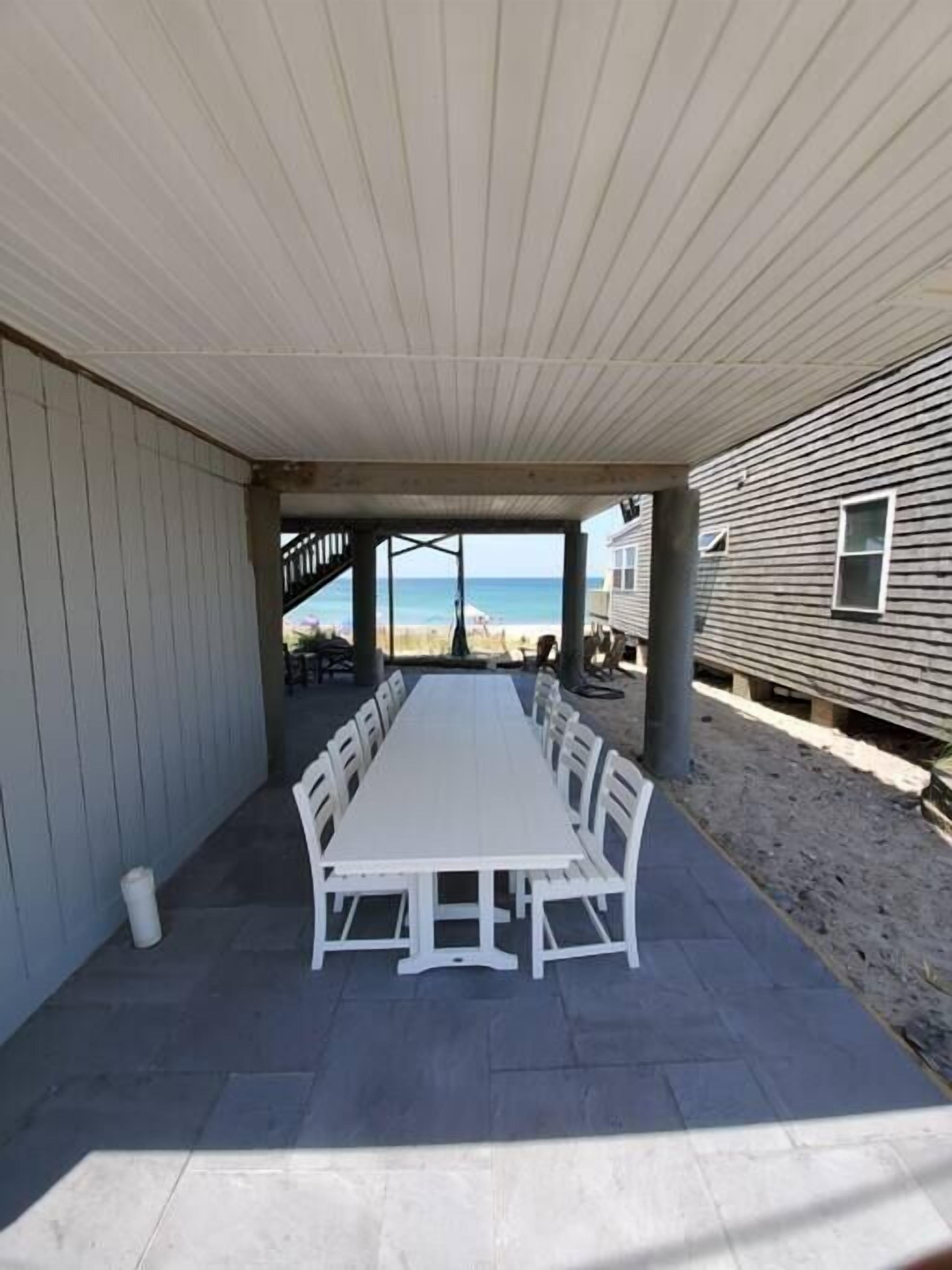 A covered patio with a long white table and chairs, overlooking a beach.