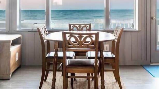 Een eetruimte met een ronde tafel en stoelen, grote ramen met uitzicht op de oceaan en een houten plafond.