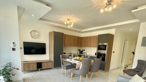 Ein modernes Wohnzimmer mit einem grauen Sofa, einem Essbereich mit Tisch für vier Personen und einer Küche im Hintergrund.