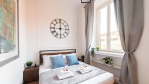Een net opgemaakt bed met een grijze deken, twee blauwe kussens en twee witte handdoeken met een afbeelding van fruit. Daarboven hangt een grote wandklok en aan de rechterkant is een raam met gordijnen te zien.
