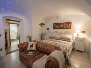 A bedroom with a bed, a brown leather sofa, a bedside table, a nightstand, and a wall-mounted shelf with heart-shaped decorations.