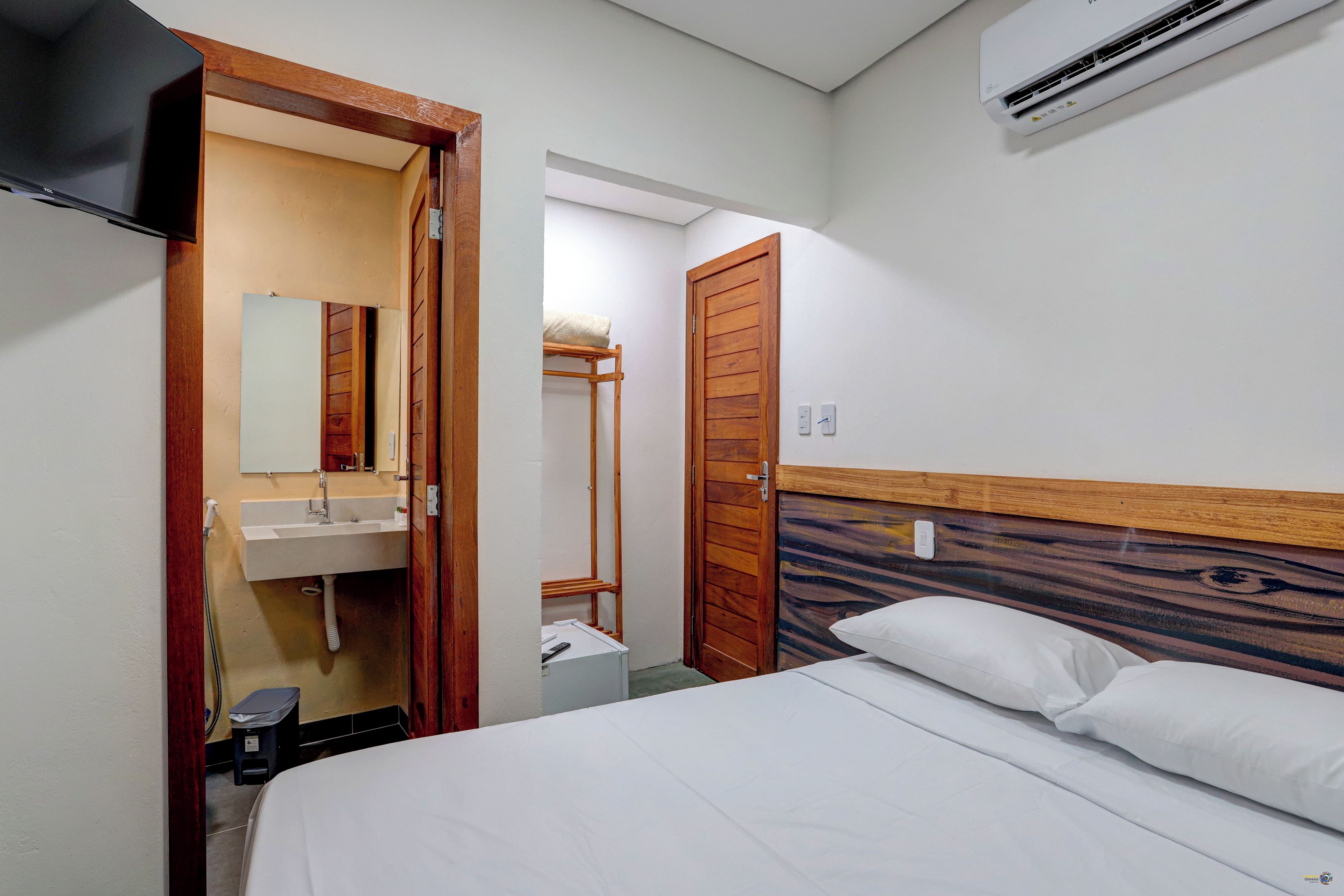 Habitación de hotel con una cama, cabecera de madera, pared blanca, puerta de madera, lavamanos blanco y un espejo.