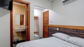 Habitación de hotel con una cama, cabecera de madera, pared blanca, puerta de madera, lavamanos blanco y un espejo.