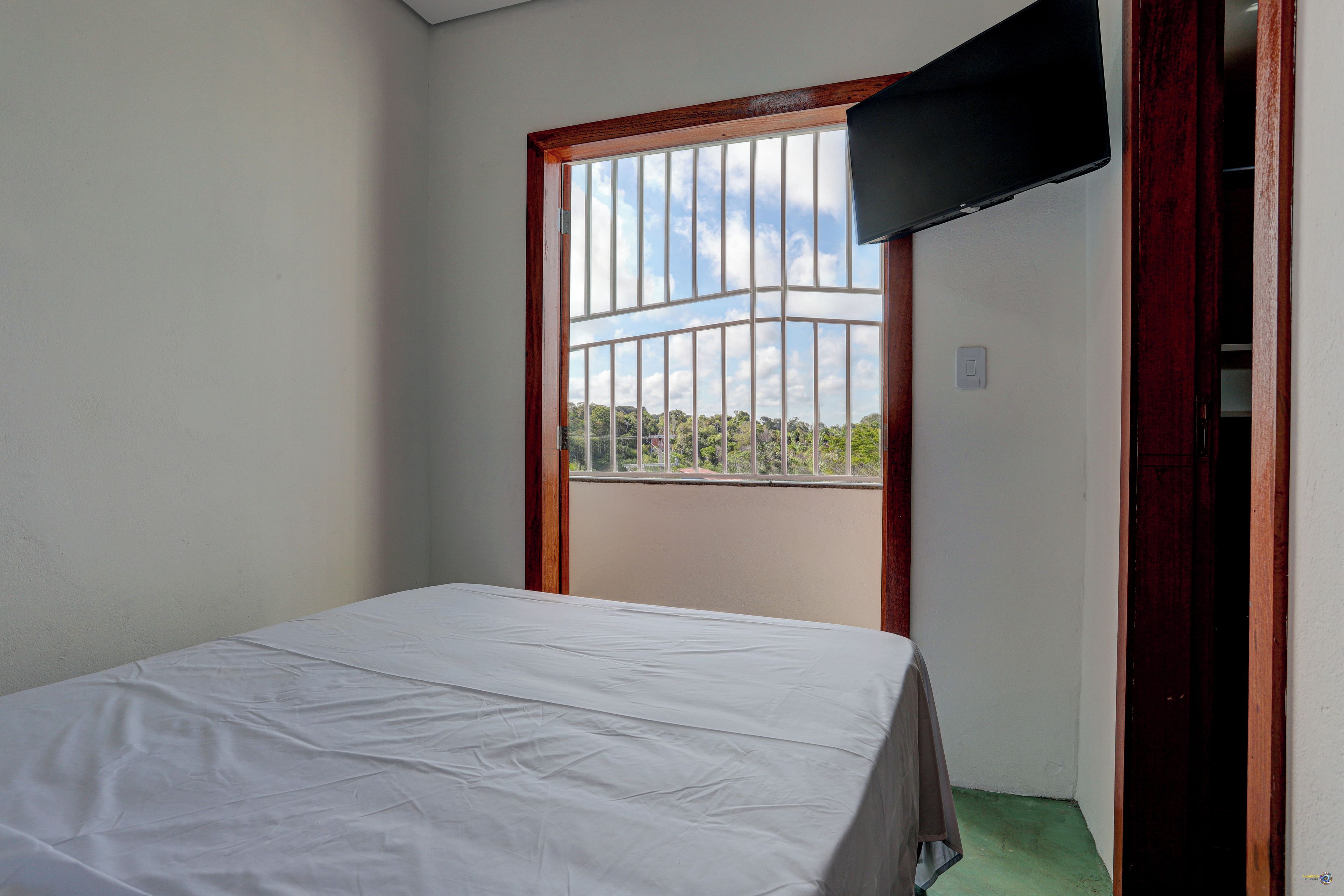 Ein Zimmer mit einem Bett, einem Fenster mit Blick auf Bäume und einem an der Wand befestigten Fernseher.