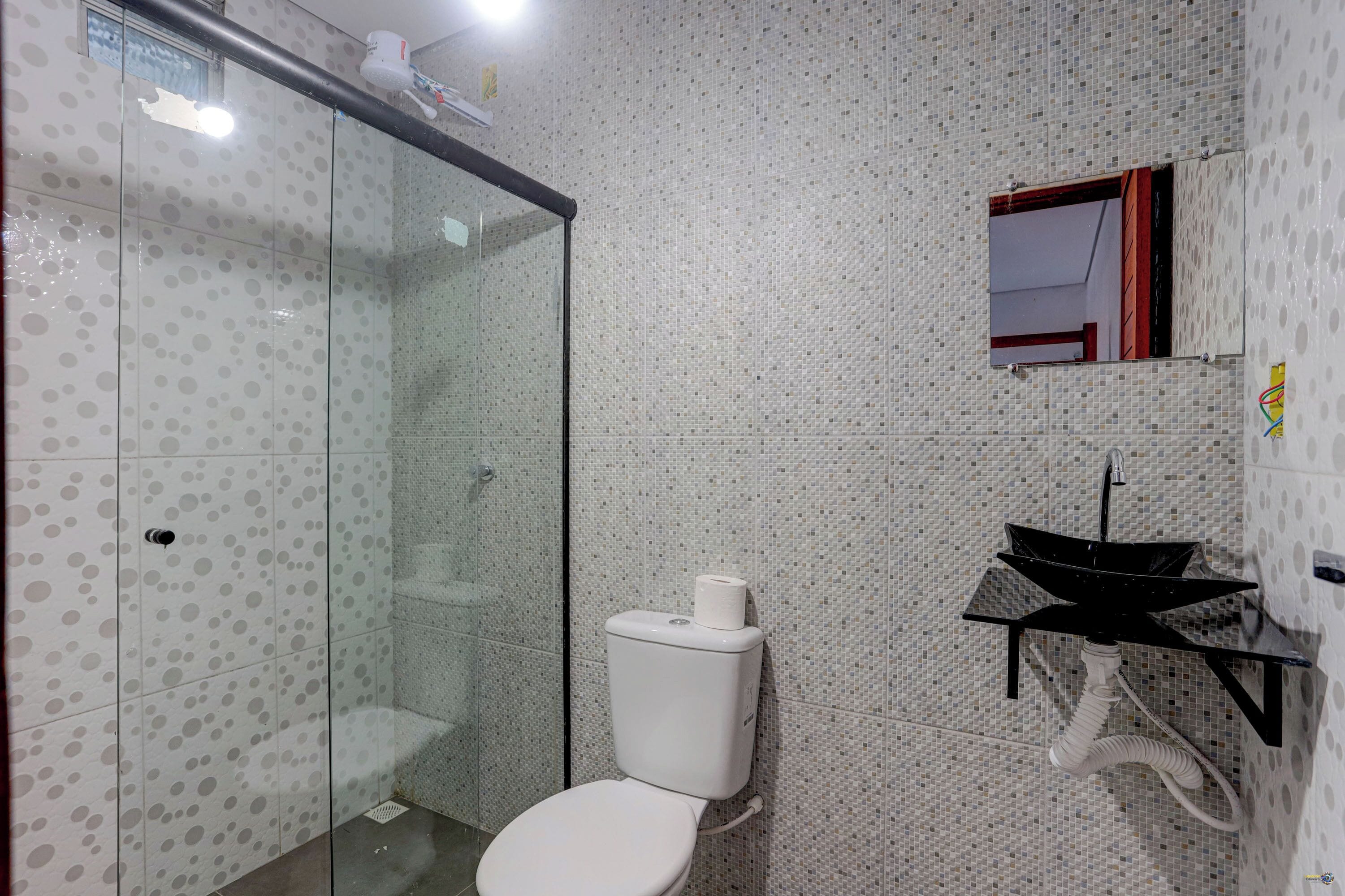 Uma casa de banho com cabine de duche em vidro, sanita, lavatório com bacia preta e espelho sobre o lavatório.