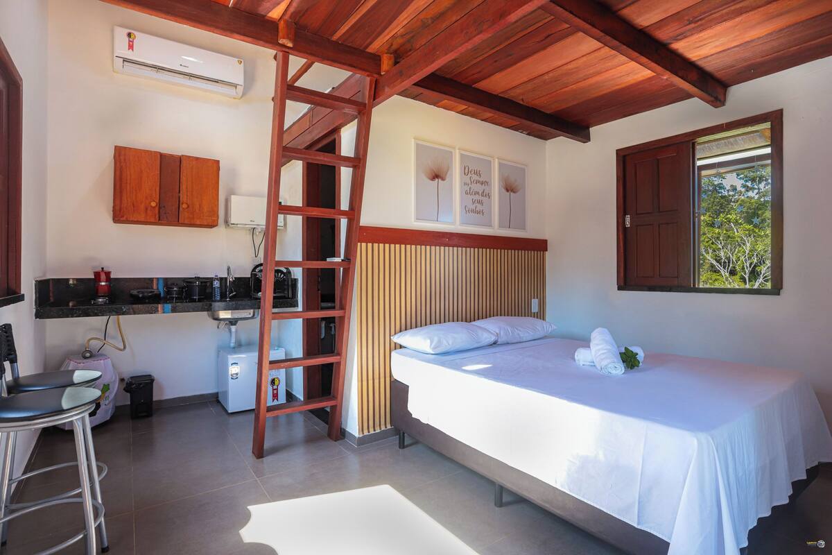 A bedroom with a bed, a wooden ladder, a kitchenette, and a window with a view.