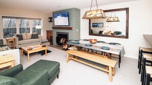 A cozy living room with a fireplace, a dining table with blue plates, and a green sofa. - 3BR/2BA, Ski in/out from A/B lift, pool & hot tub (Ludlow)