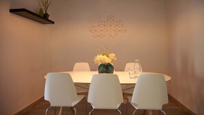 Un comedor con una mesa blanca, tres sillas blancas, un florero con flores, un adorno mural geométrico y una lámpara colgante.
