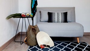 Una sala de estar moderna con un sofá gris, almohadones estampados, una mesita de noche con un sombrero y gafas de sol, y una planta decorativa.
