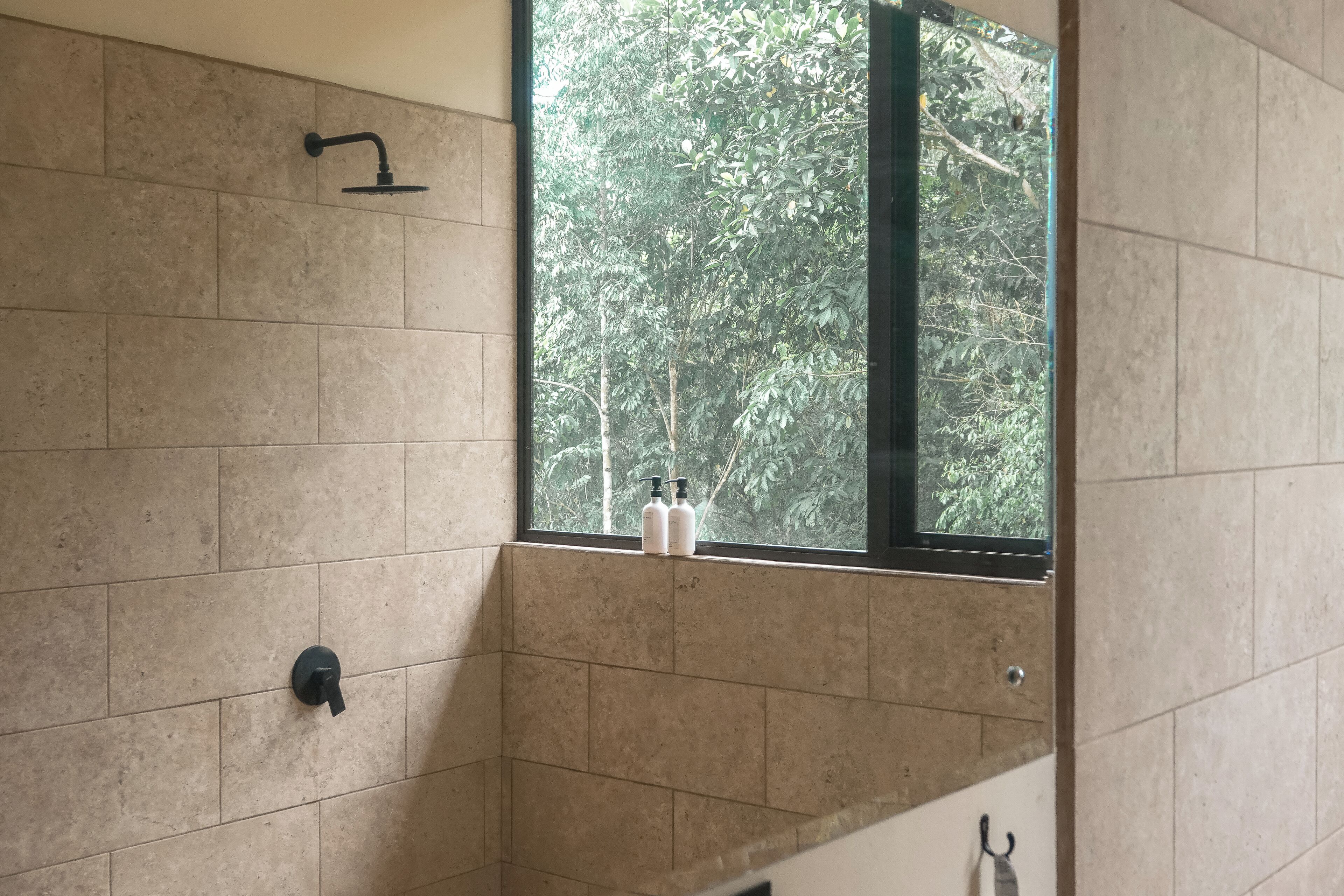 A bathroom with beige tiles, a window with a view of trees, and two bottles on the window sill.