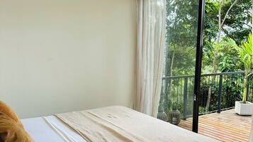 A bedroom with a bed, a window with sheer curtains, and a view of a wooden deck and greenery outside.